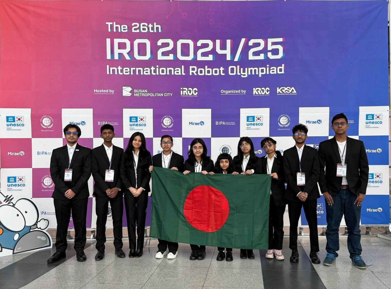 Group photo of the Bangladesh team at the 26th International Robot Olympiad, proudly holding the national flag in front of the event banner.